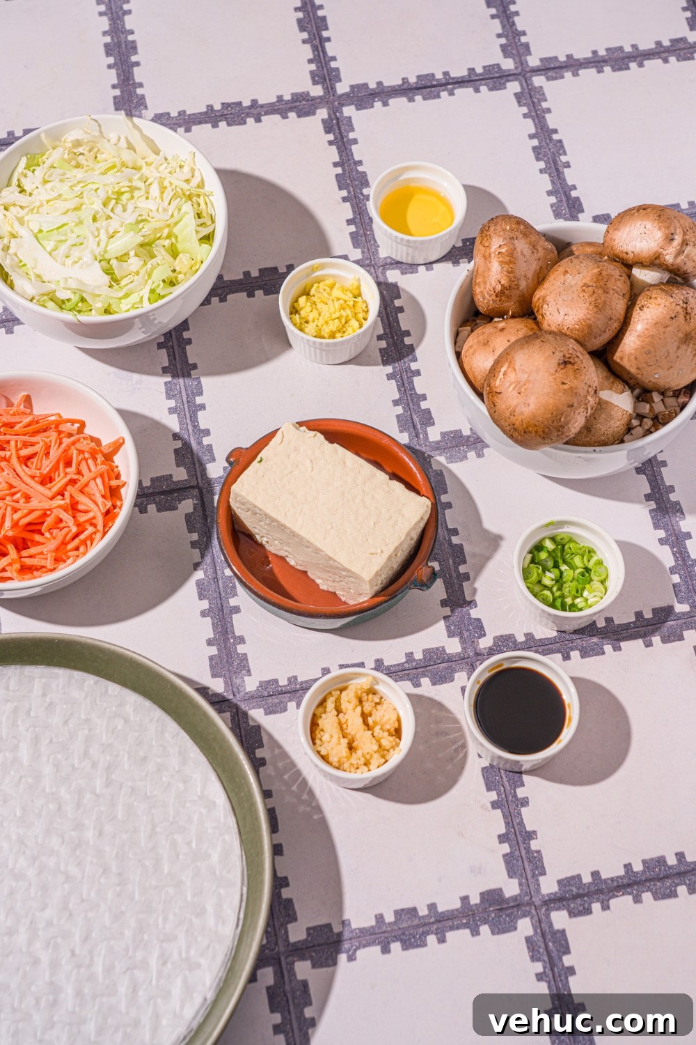 Crispy Rice Paper Veggie Dumplings: Viral TikTok Recipe 4 A tiled counter with several bowls in various sizes containing ingredients to make vegetable rice paper dumplings including sheets of rice paper, tofu, shredded cabbage, shredded carrots, mushrooms, garlic, sliced green onions, and more.