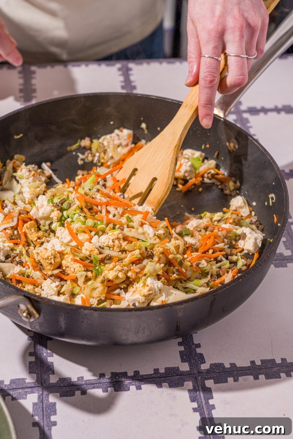 Crispy Rice Paper Veggie Dumplings: Viral TikTok Recipe 7 A skillet with a wooden spoon cooking a vegetable mixture including diced mushrooms and shaved carrots and cabbage. The skillet is on a tiled counter.